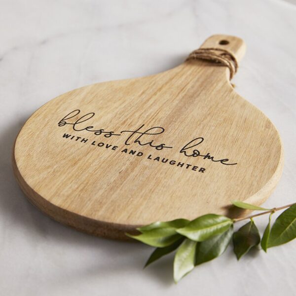 A wooden cutting board with the words " bless this home."