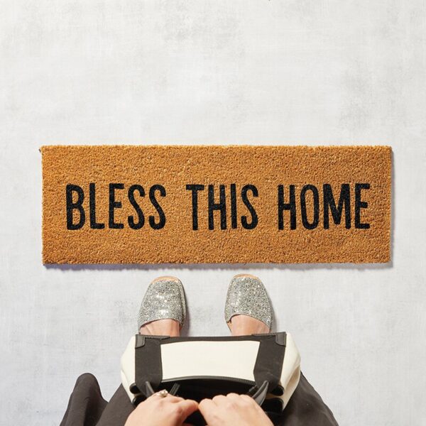 A person is holding their phone in front of a sign that says " bless this home ".